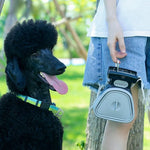 Black dog with a colorful collar standing next to a person holding a blue pet waste scooper in a park setting.