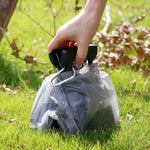 Hand holding a black pet waste scooper wrapped in a plastic bag with a clip on grass